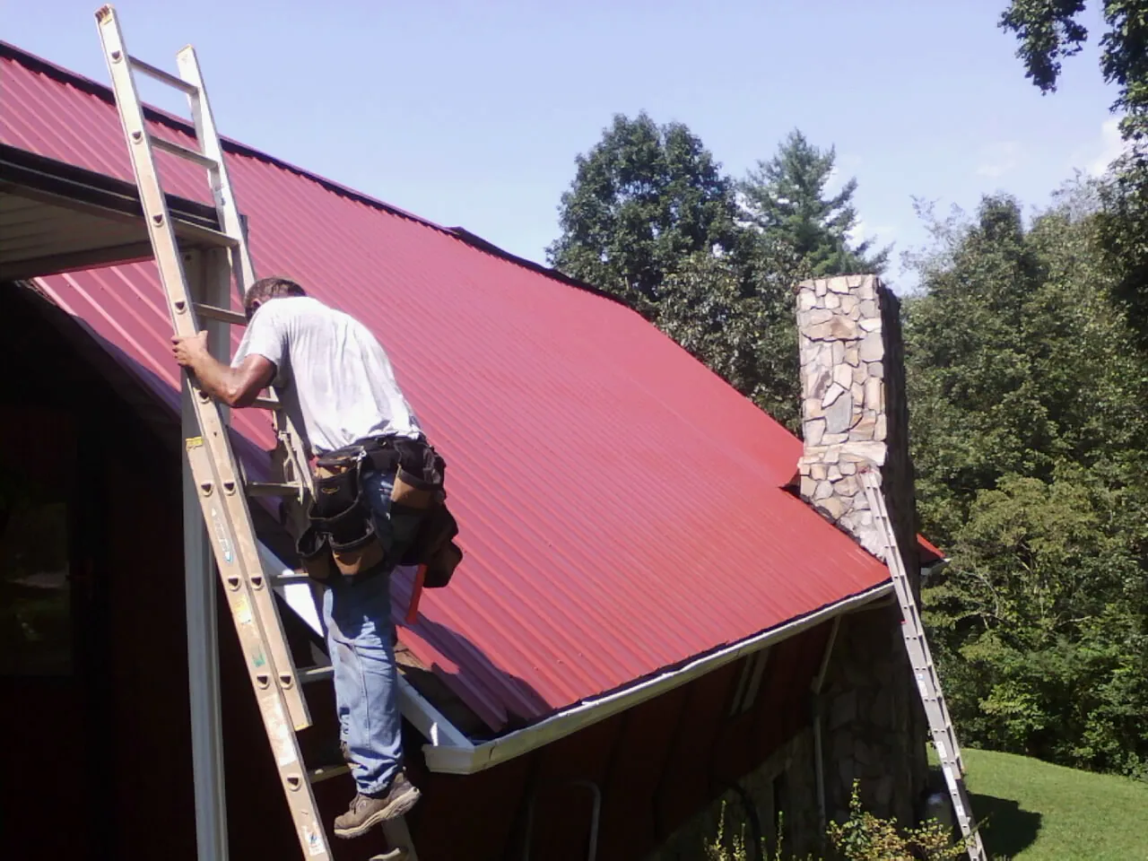 Roofer on a ladder beside a red metal roof and stone chimney — Soffit & Fascia Repair by Roofing Brothers in Good Hope
